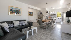 Un salon moderne avec un canapé en cuir noir, une table à manger avec des chaises et un coin cuisine doté de meubles blancs et d’un grand téléviseur.