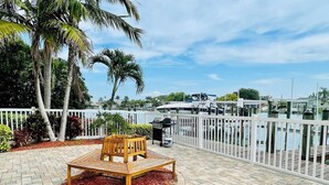 A wooden bench in a paved outdoor area with a white fence and palm trees in the background.