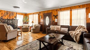 A cozy living room with wooden walls, a stone fireplace, and a variety of seating including sofas and armchairs. - A rustic retreat nestled in the heart of Boulder Bay (Big Bear Lake)