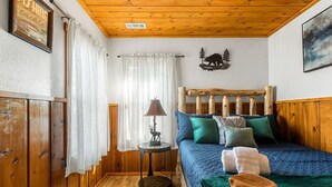A cozy room with a wooden ceiling, a bed with blue bedding, a small table with a lamp, and a window with white curtains. - A rustic retreat nestled in the heart of Boulder Bay (Big Bear Lake)