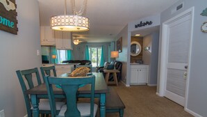 A dining area with a table and chairs, a kitchen in the background, and a door with a 'Welcome' sign. - Seaside Escape in Pebble Beach  (Emerald Isle)