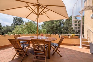 Una mesa y sillas de madera bajo un paraguas grande en una terraza con vistas a árboles y una escalera en espiral blanca.