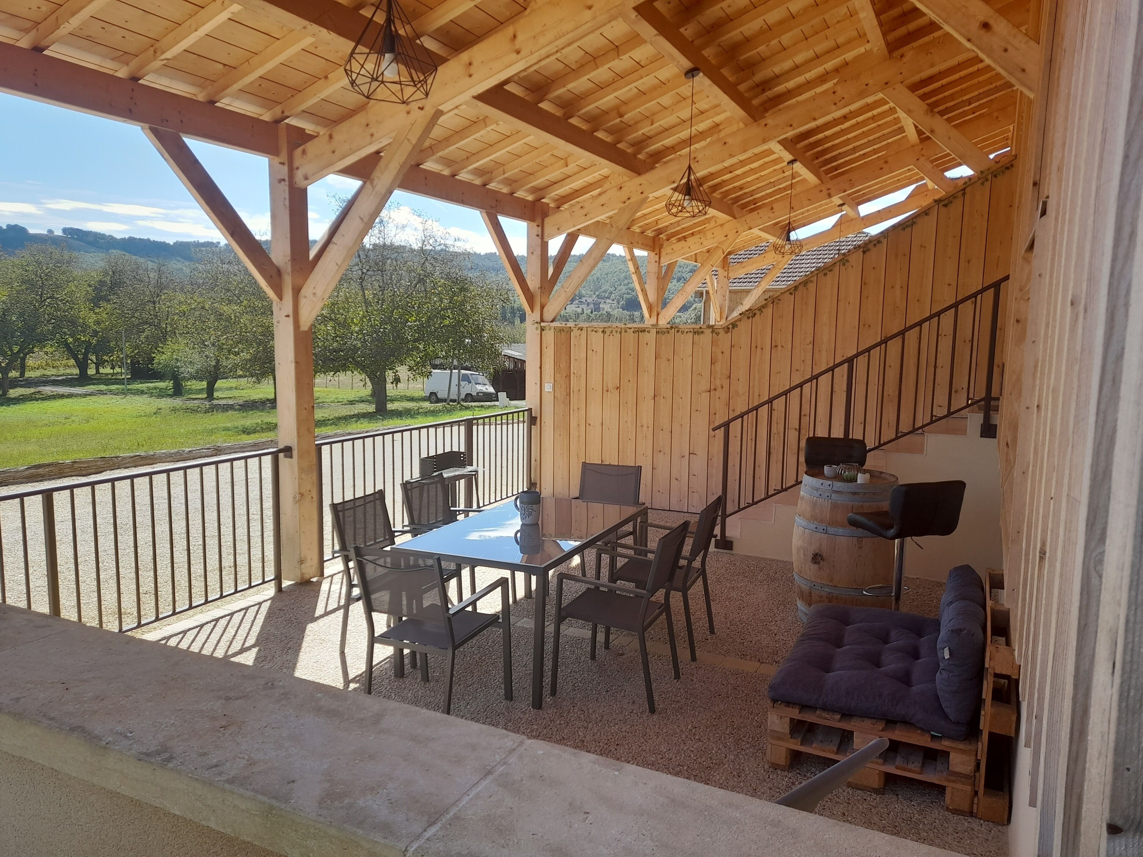 Una terraza de madera con una mesa, sillas, un barril y vistas a un paisaje verde.