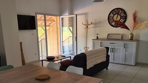 Un salon moderne avec une table à manger en bois, des chaises blanches, un canapé noir, un téléviseur fixé au mur et une grande fenêtre donnant sur un paysage verdoyant.