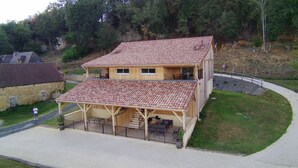 Une maison en bois à deux étages, avec un toit en tuiles, une terrasse couverte dotée de meubles d’extérieur et une allée pavée.