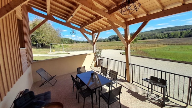 Un espace extérieur aménagé, abrité, avec une table et des chaises, un petit foyer et une vue sur un champ et des collines.