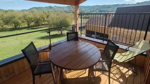 A wooden deck with a round table and chairs, overlooking a green landscape with trees and hills.