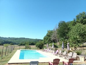 Piscina retangular com espreguiçadeiras e guarda-sóis, cercada por um espaço vedado e árvores.