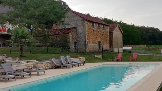 A rectangular swimming pool with lounge chairs and a covered area, surrounded by a stone building and greenery.
