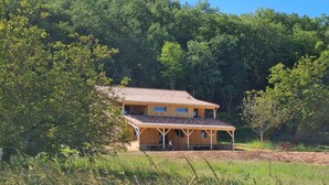 A two-story wooden house with a tiled roof, surrounded by dense greenery and trees.