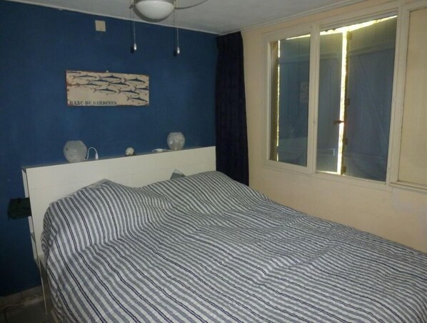 Un dormitorio con colcha a rayas, cabecera, lámpara de pared y ventana con cortinas.