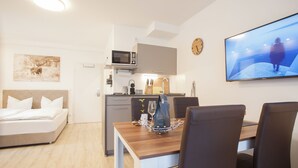 Una habitación de hotel moderna con cama, una mesa de comedor con sillas, una zona de cocina, un televisor de pantalla plana y un reloj en la pared.