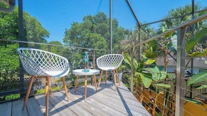 Una terraza de madera con dos sillas blancas y una mesita, rodeada por una valla de metal y abundante vegetación.