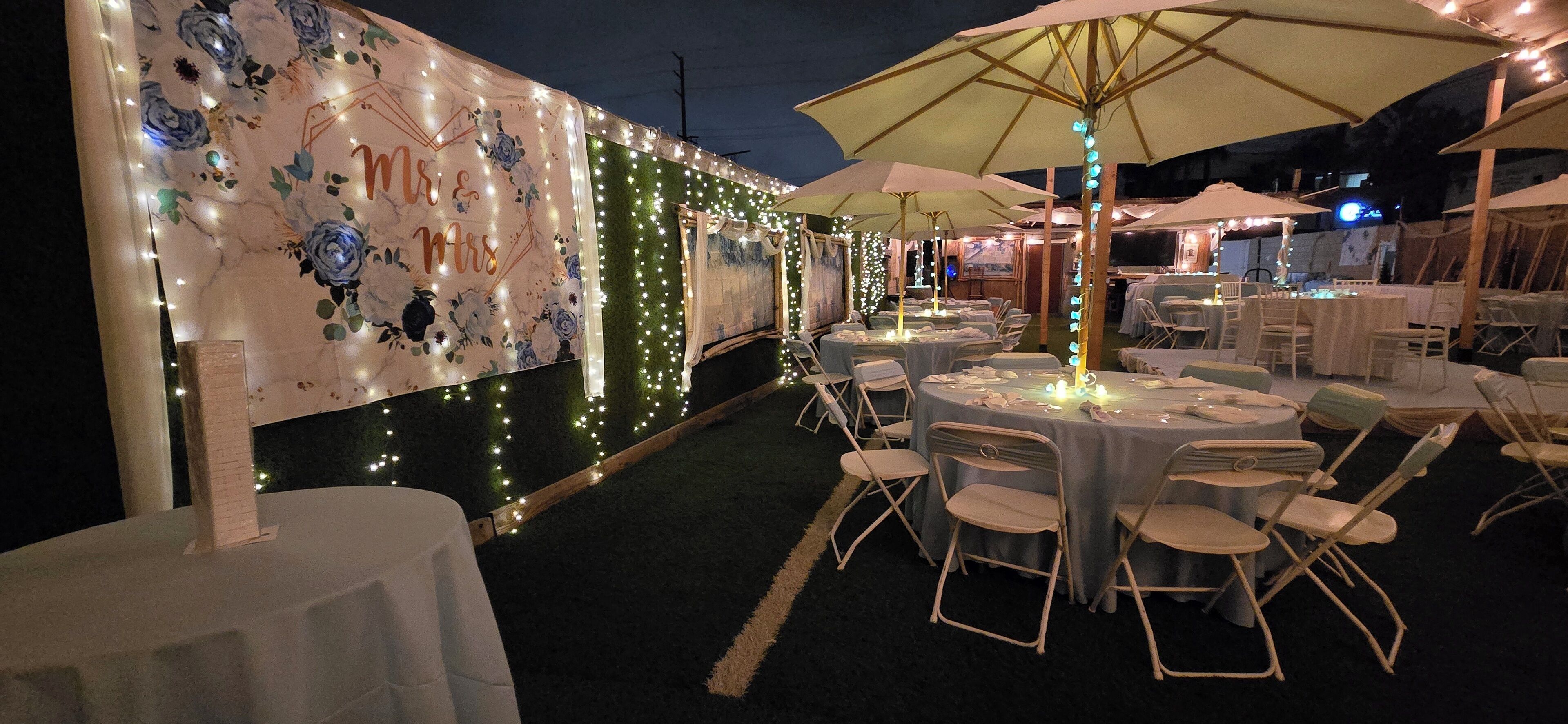 Un espace extérieur aménagé pour les repas, avec des tables et des chaises rondes, décoré de guirlandes lumineuses et de parasols.