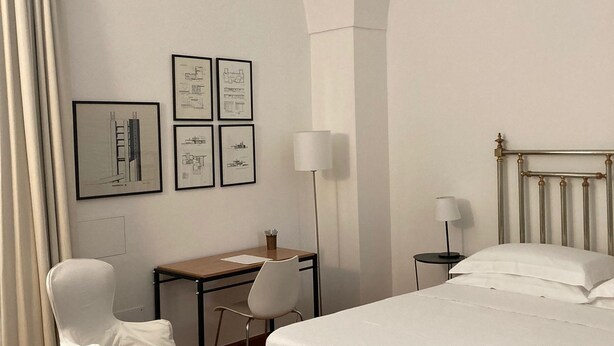 Habitación de hotel con cama, escritorio, dos sillas, lámpara y dibujos arquitectónicos enmarcados en la pared.