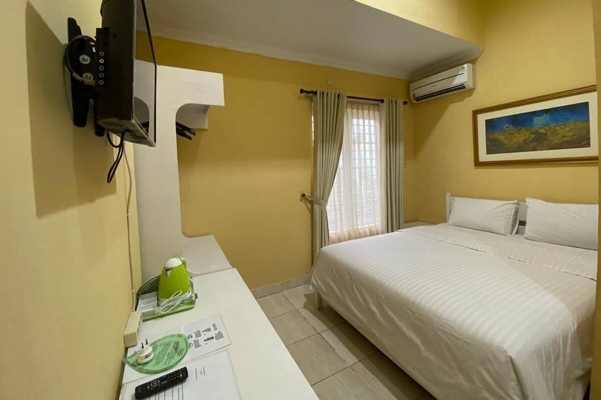 Une chambre d’hôtel avec un lit, un bureau avec un téléphone et un objet vert, un tableau au mur et un climatiseur.