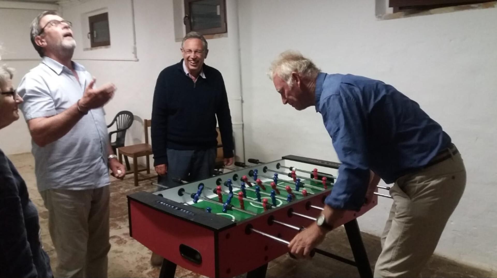 Un groupe de personnes joue au baby-foot dans une pièce à l’aménagement simple, comprenant une table et des chaises.