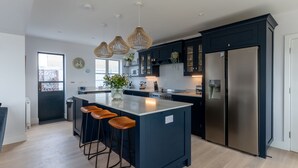 A modern kitchen with dark cabinetry, a large island with stools, stainless steel appliances, and pendant lighting. - The Old Coastguards, Sleeps 8, Pets, Hot Tub (Braunton)