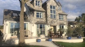 A two-story house with a stone facade, large windows, and a covered patio area with outdoor seating. - The Old Coastguards, Sleeps 8, Pets, Hot Tub (Braunton)