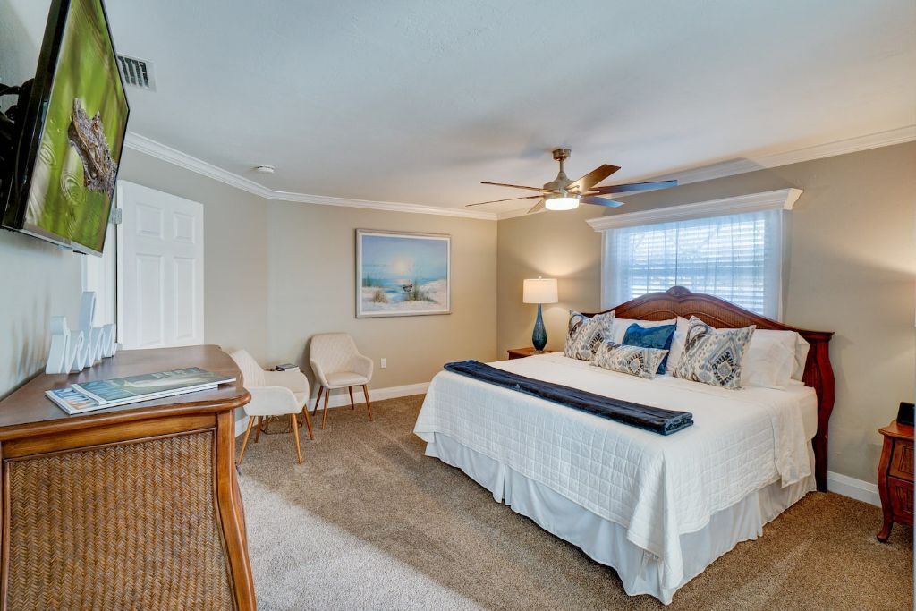 A bedroom with a bed, a desk, a chair, a ceiling fan, a window with blinds, and a painting on the wall.
