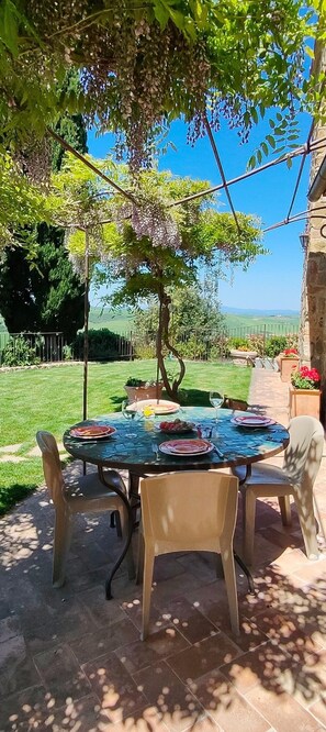 Un área de comedor al aire libre con una mesa preparada para dos personas, rodeada de sillas, bajo una pérgola con enredaderas colgantes.
