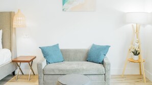 A modern room with a grey sofa, two blue cushions, a small wooden table, a floor lamp, and a painting of a beach scene. - SKS - Superior Room, 1 King Bed with Sofa bed, Non Smoking (Long Beach)