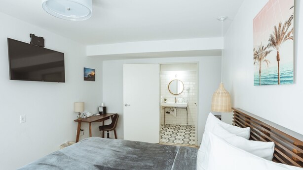 A bedroom with a bed, a desk, a chair, a TV mounted on the wall, a painting, and a bathroom visible through an open door. - ADA - Deluxe Room, 1 Queen Bed, Accessible Non Smoking (Long Beach)