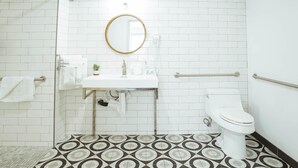 A bathroom with a white tiled wall, a round mirror, a white sink, a toilet, and a patterned floor. - ADA - Deluxe Room, 1 Queen Bed, Accessible Non Smoking (Long Beach)