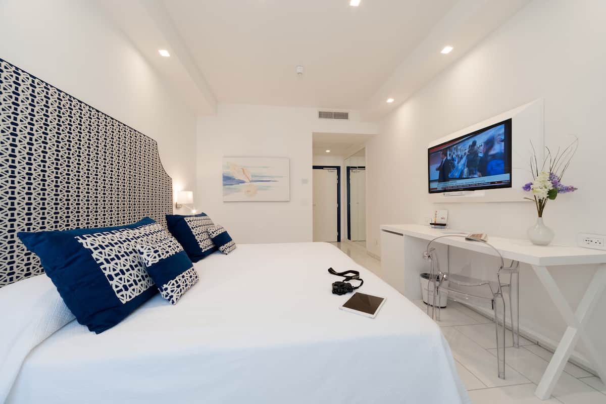Ein modernes Hotelzimmer mit einem ordentlich bezogenen Bett, einem Schreibtisch mit Computer und einem an der Wand montierten Fernseher.