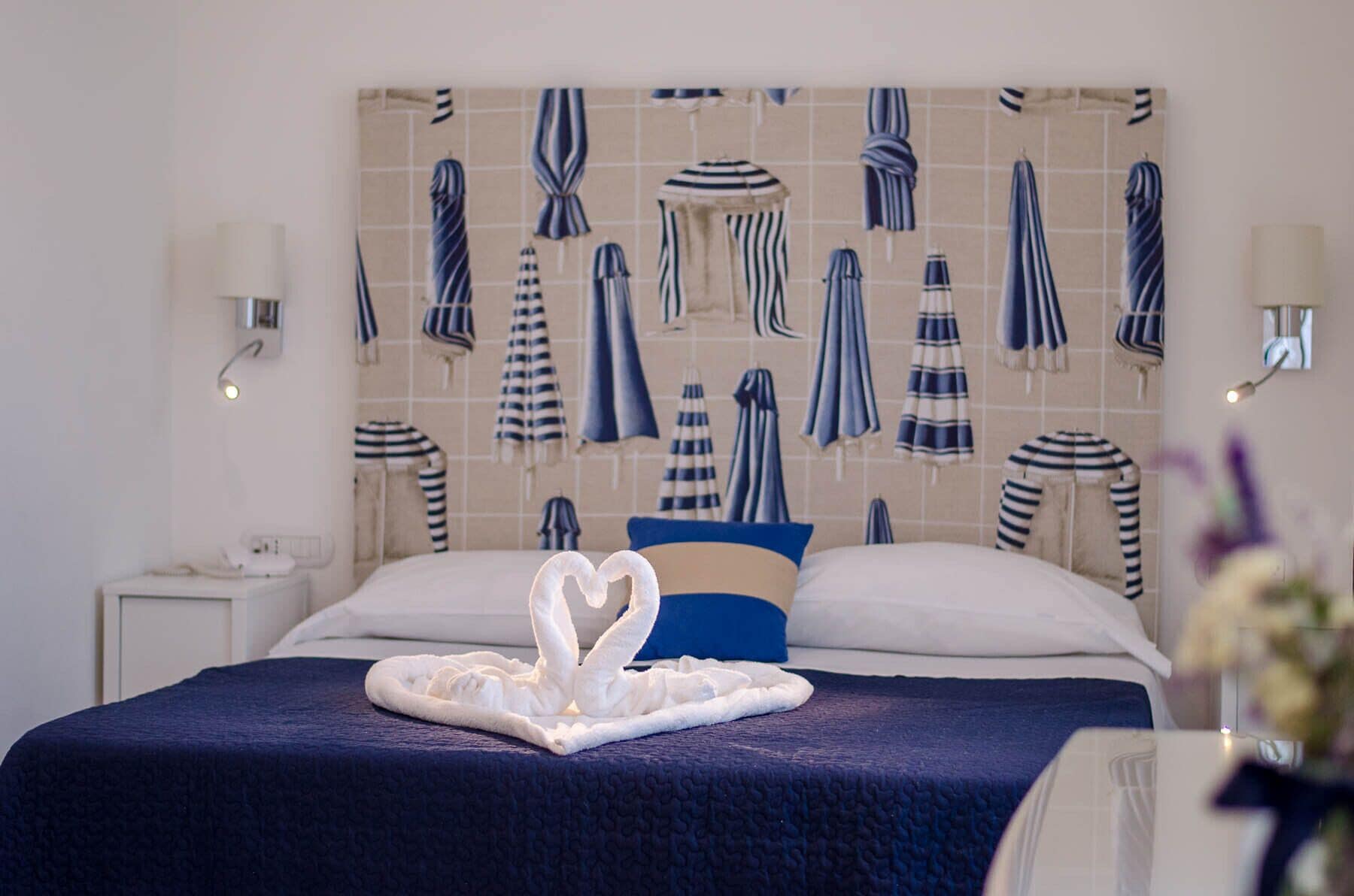 Habitació d'hotel amb un llit, una manta blava, una tovallola en forma d'un cigne blanc i una paret decorada amb tovalloles amb ratlles.