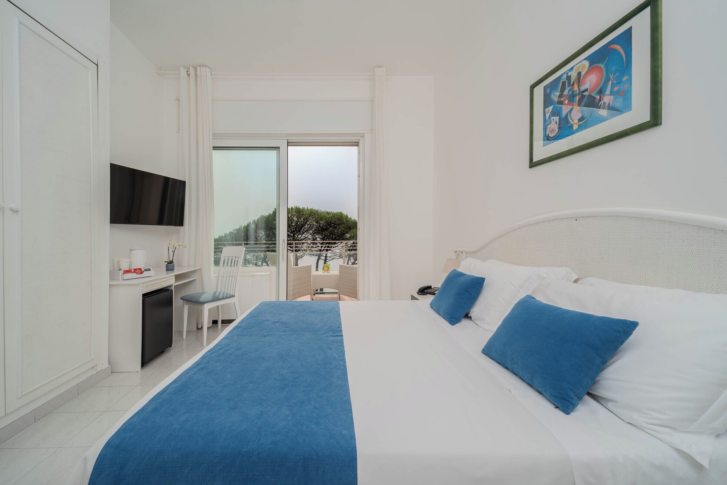 A hotel room with a bed, a desk with a TV, a chair, and a balcony with a view.