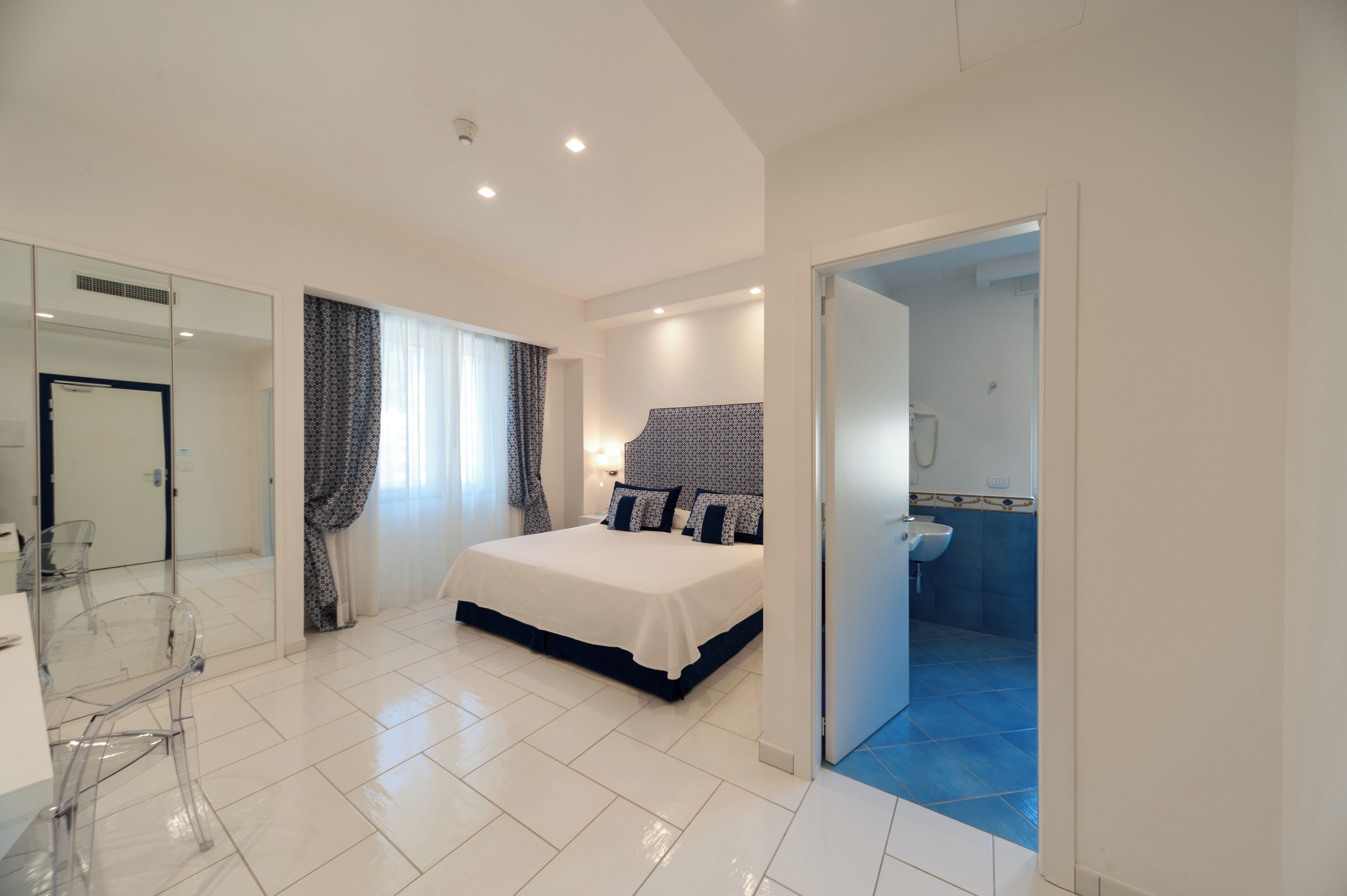 A modern bedroom with a large bed, a glass desk, and a bathroom with blue tiles.