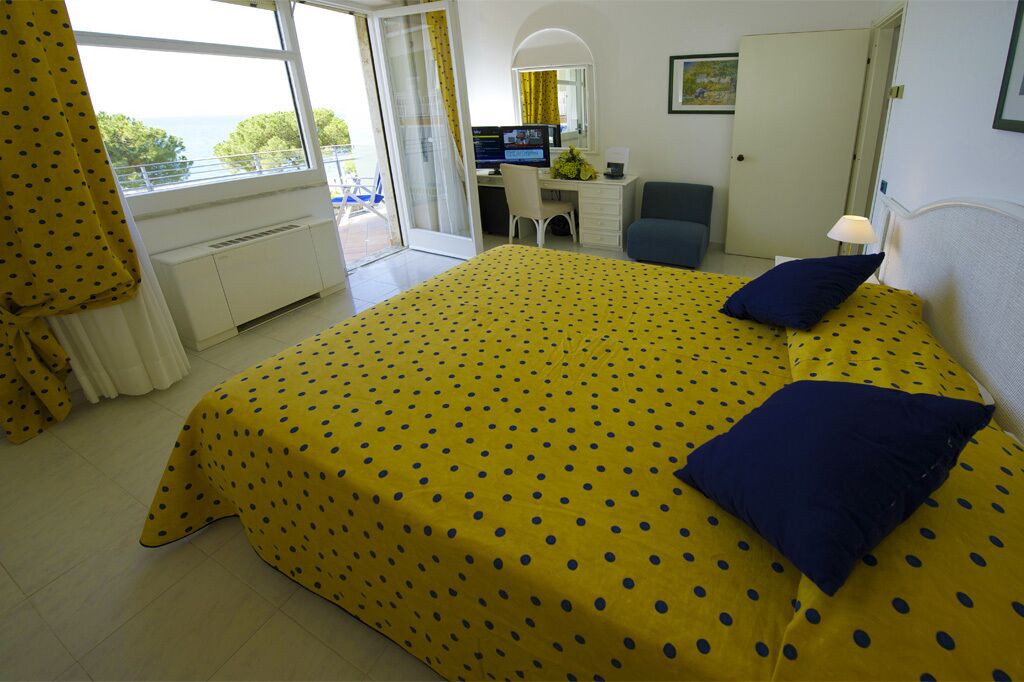 A hotel room with a bed, a desk, a chair, a computer, and a view of the sea.