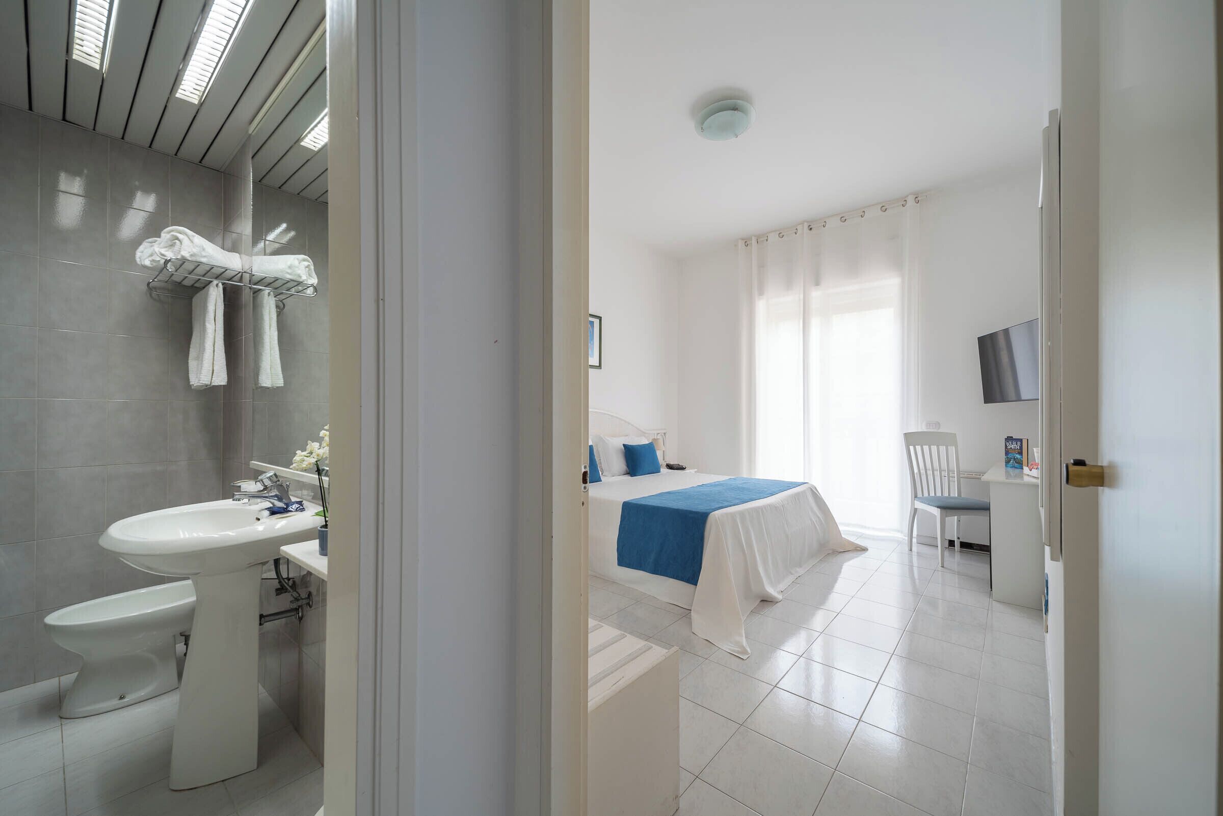 A hotel room with a bathroom, a bed with a blue bedspread, a desk, and a chair.