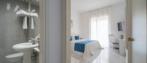 A hotel room with a bathroom, a bed with a blue bedspread, a desk, and a chair.