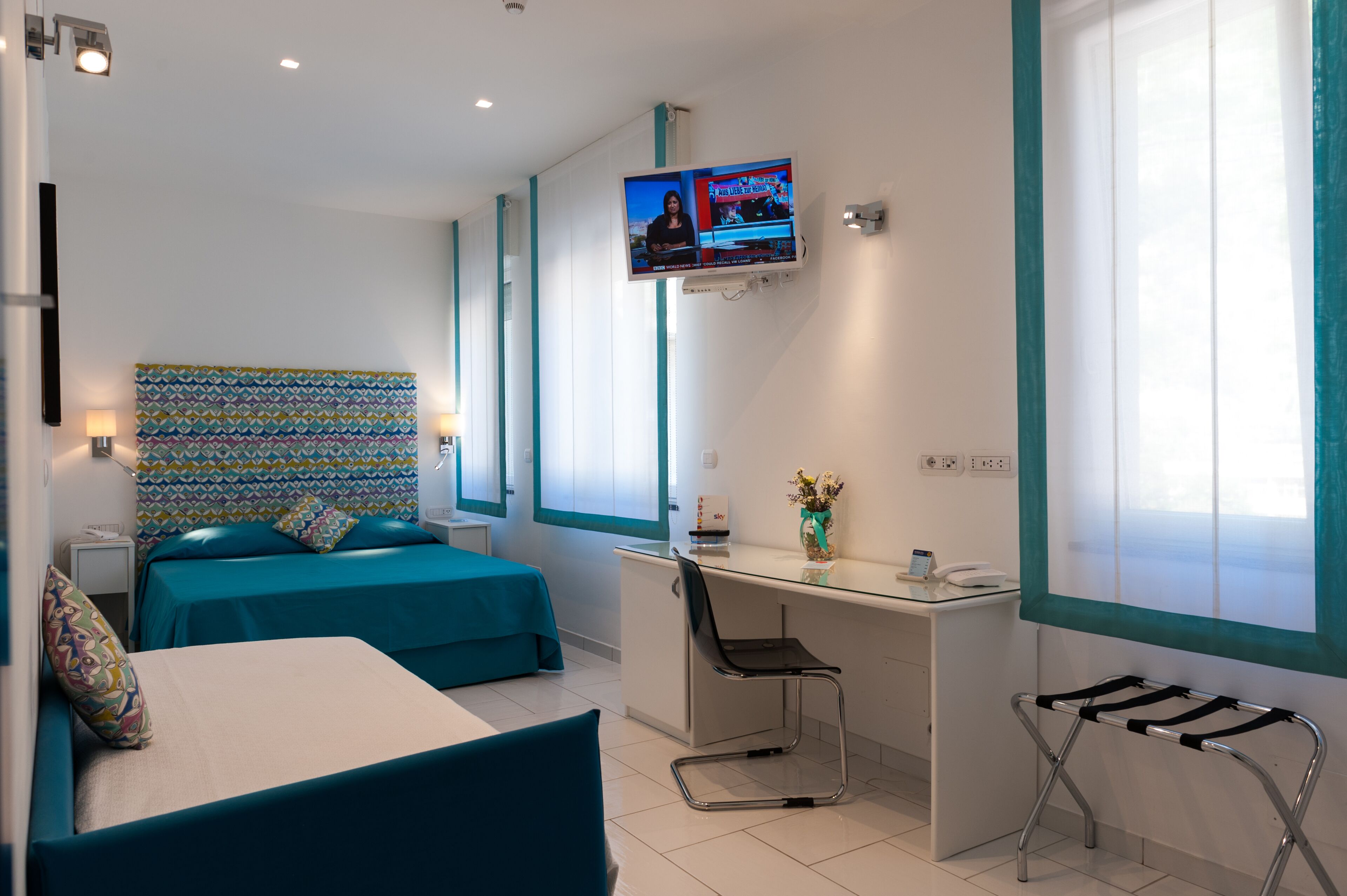 A hotel room with two beds, a desk, a chair, a TV, and a window.