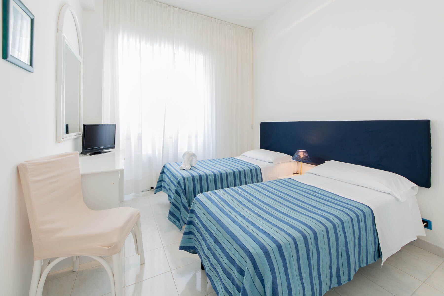 A hotel room with two beds, a chair, a TV, and a window with curtains.