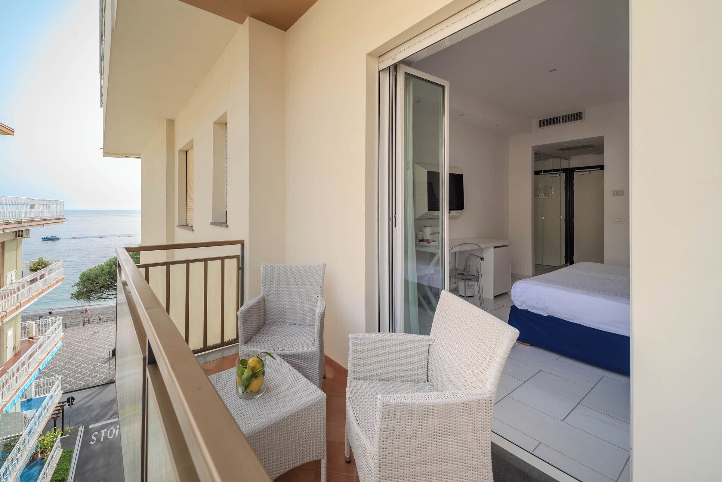 Ein Balkon mit zwei Korbstühlen, einem kleinen Tisch und einer Obstschale, mit Blick aufs Meer.