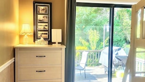 A bedroom with a wooden dresser, a bed, a window with a view of the outdoors, and a blue mat at the entrance.