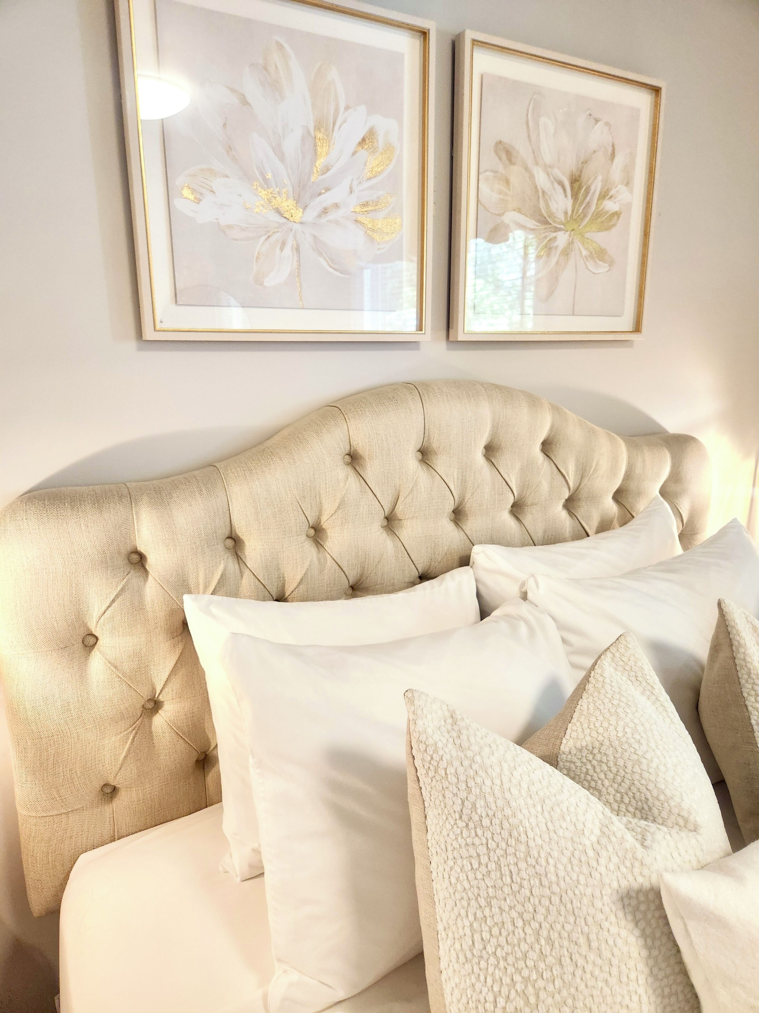Un dormitorio con cabecera capitonada, ropa de cama blanca y dos cuadros con motivos florales en la pared.