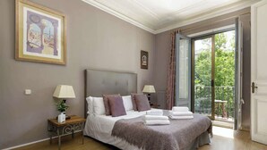 Ein Schlafzimmer mit einem großen Bett, zwei Nachttischen mit Lampen, einem gerahmten Bild an der Wand und einem Balkon mit Blick auf Grünflächen.