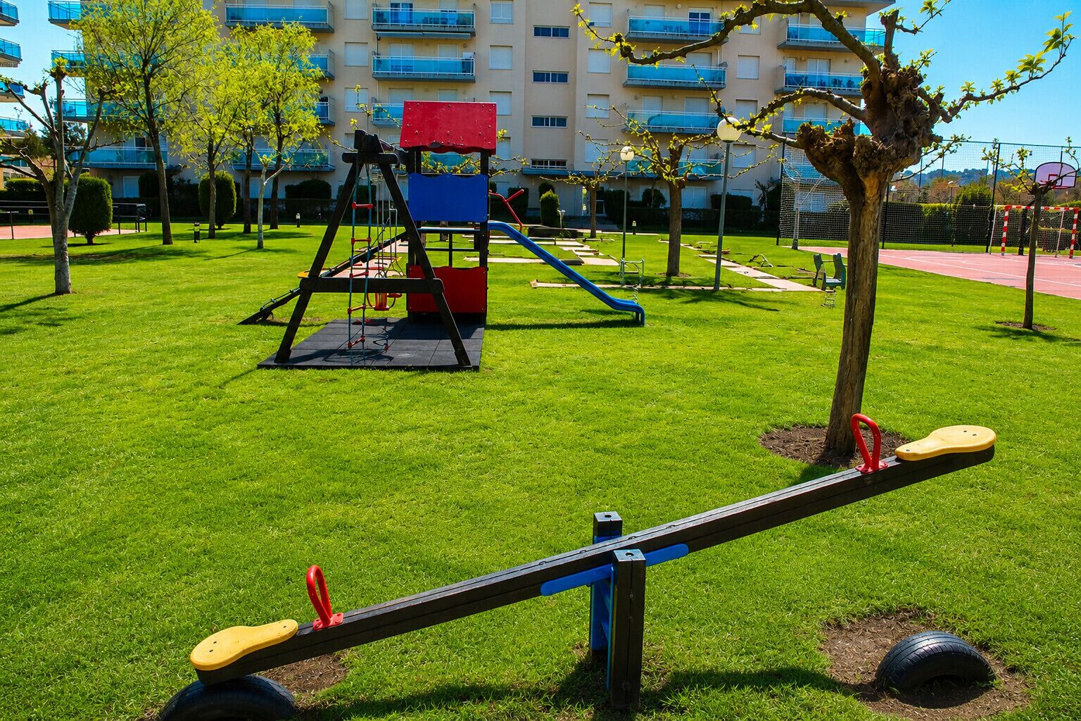 Un parque infantil con sube y bajas y columpios, rodeado de césped y árboles, con un edificio al fondo.