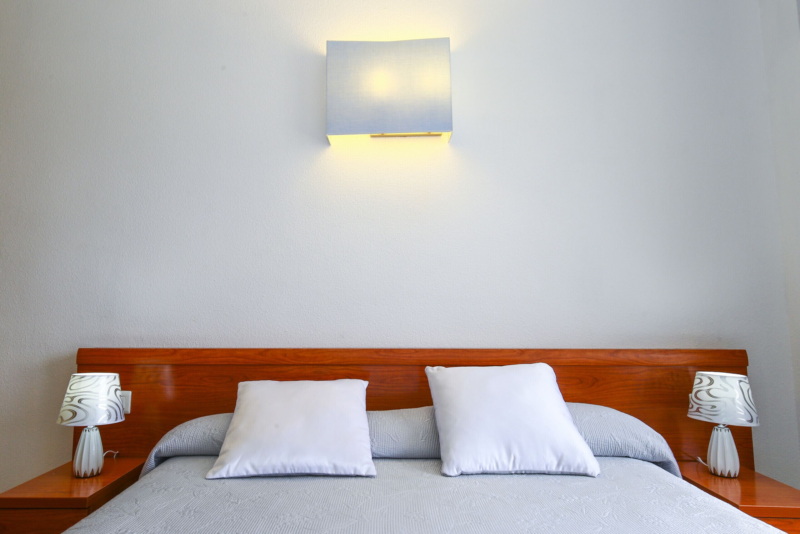 Cama bem arrumada com duas almofadas, cabeceira de madeira e duas luminárias laterais.