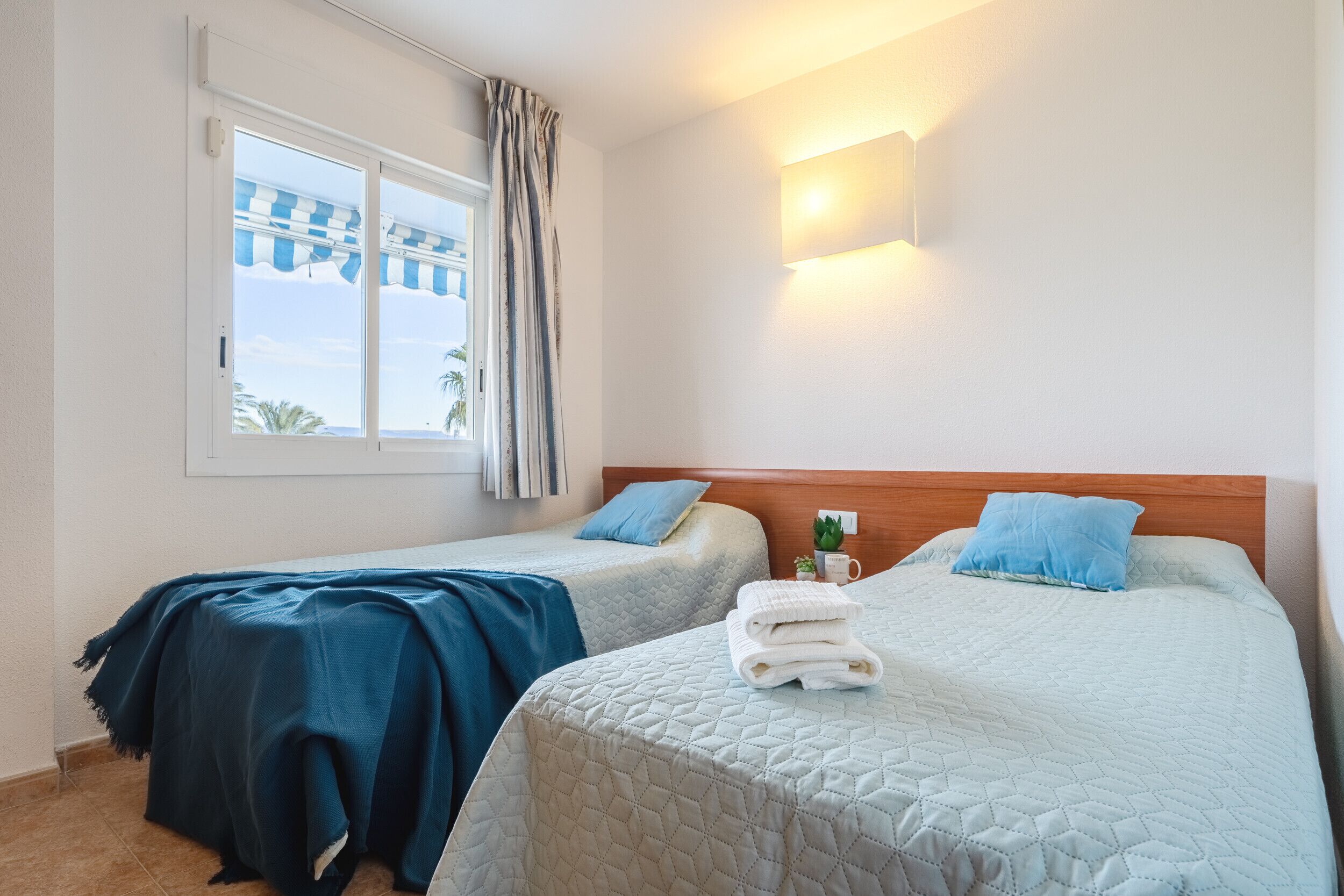 A hotel room with two beds, a window with a view, a wall-mounted light, and a small plant on the nightstand.