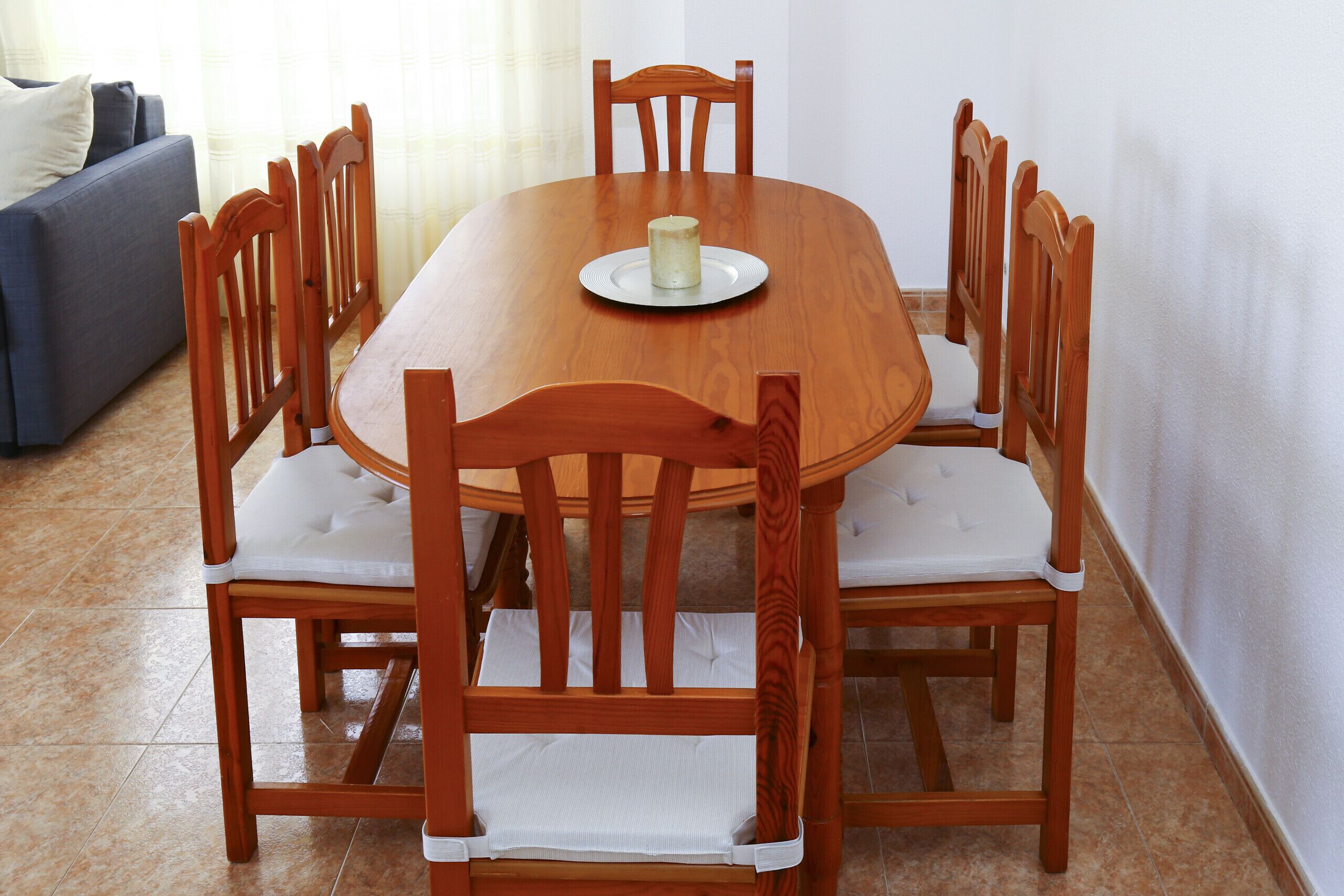 A wooden dining table with six chairs, a white plate with a candle, and a grey sofa in the background.