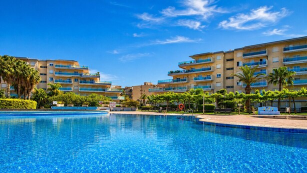 A swimming pool with clear blue water, surrounded by multi-story buildings with balconies, palm trees, and a clear sky. - Residencial LOS JUNCOS III (La Pineda)