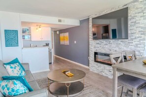 A modern living room with a round coffee table, a sofa with patterned cushions, a dining area with a bench, and a kitchen in the background.