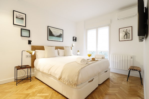 A bedroom with a large bed, a bedside table with a lamp, a window with a view, and framed pictures on the wall.