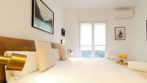 A hotel room with a large bed, a headboard, a window with a view, a wall-mounted air conditioner, and a framed picture on the wall.