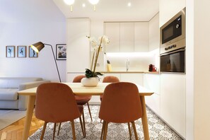 Eine moderne Küche mit weißen Schränken, einem Holztisch und orangefarbenen Stühlen. Auf dem Tisch steht ein Topfpflanze und darüber befindet sich eine Mikrowelle.